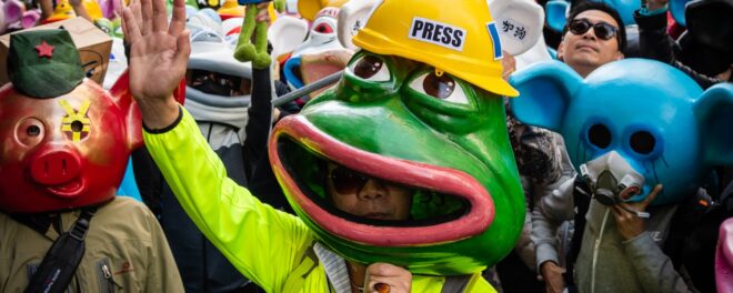En person iklädd en stor Pepe the Frog-mask med gul hjälm märkt "PRESS" håller upp en hand i en folkmassa, troligen vid en demonstration.