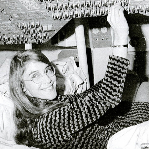 Svartvit bild av en glad kvinna som ligger på rygg och arbetar med kablar och utrustning ovanför sig.