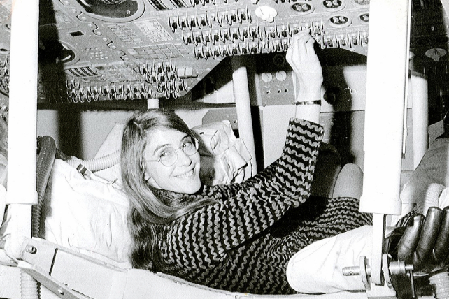Svartvit bild av en glad kvinna som ligger på rygg och arbetar med kablar och utrustning ovanför sig.