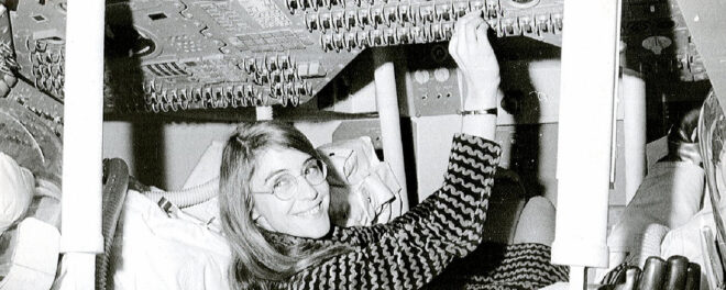 Svartvit bild av en glad kvinna som ligger på rygg och arbetar med kablar och utrustning ovanför sig.