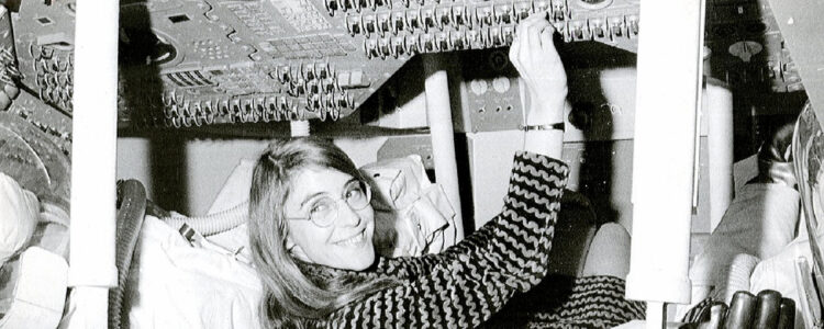 Svartvit bild av en glad kvinna som ligger på rygg och arbetar med kablar och utrustning ovanför sig.