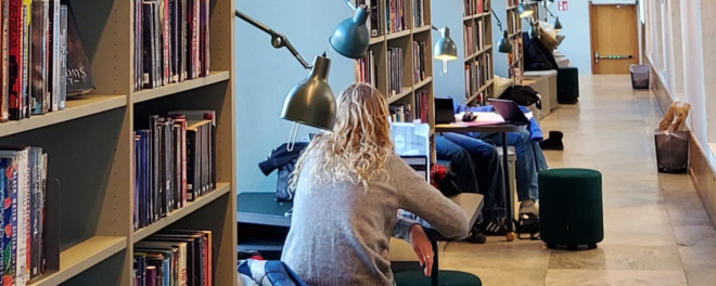 Blond tjej sitter med ryggen mot kameran och läser på sin dator inne på biblioteket.