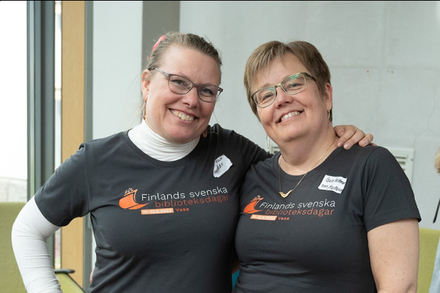 På bilden syns ett porträtt i närbild av två leende kvinnor. Båda bär glasögon och bär en svart t-shirt. På tröjan står det Finlands svenska biblioteksförening, Vasa.