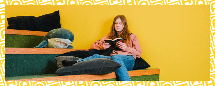 Flicka sitter och läser på inomhustrappa som fungerar som sittplats. Kuddar och nallehaj. Gul vägg.