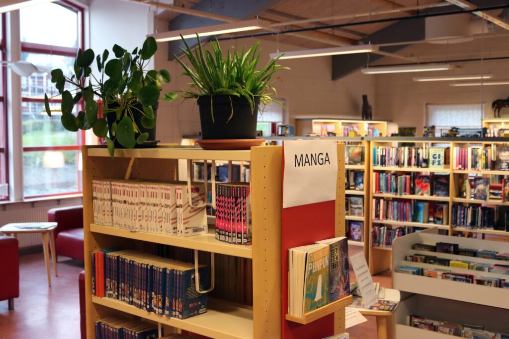 Färgglatt fotografi på bokhyllor med böcker i. Ovanpå en bokhylla är gröna växter och på kanten sitter ett papper med texten "MANGA"