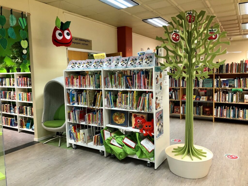 Bokhylla med många böcker i. Ovanför hänger en tyggubbe i form av ett äpple och bredvid hyllan står en möbel formad som ett träd. 