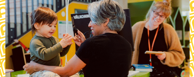 Litet barn sin sitter på ett bord i höjd med en stående vuxen vinna. Kvinnan håller i barnet ler. Barnet och kvinnan håller i varsin sak.