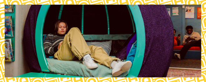 Ung person sitter i en fåtölj med hörlurar på huvudet. Det ena benet är uppdraget.