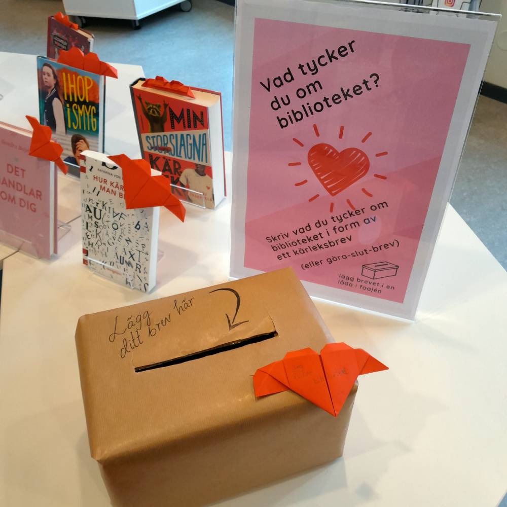 Rosa affisch med text "Vad tycker du om biblioteket?" på ett bord. Intill en brun låda med ett avlångt hål för att lägga i brev.