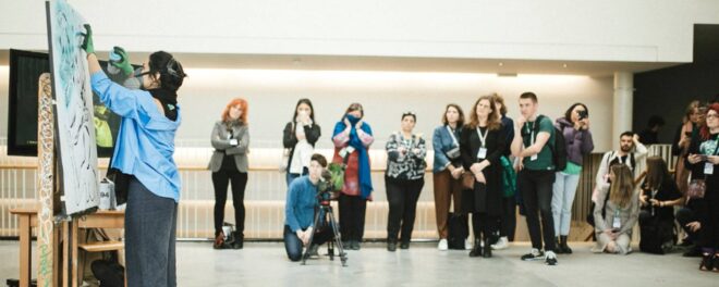 Ett staffli och en konstnär som målar till vänster i bild. I bakgrunden ett tiotal människor som följer målandet.