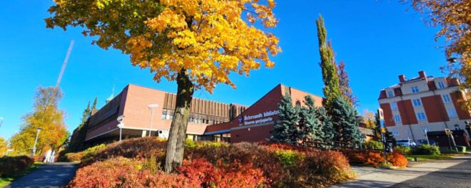 En röd byggnad och några stadshus till höger i bild. En klarblå himmel ovanför, och i förgrunden ett stort träd med gula, skinande löv. Längst ner en växtlighet i form av låga buskar med röda blad.