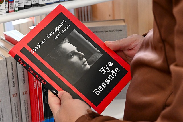 Händer håller boken 'Nya Resande' av Stephan Skougaard Carlsson med rött omslag och svartvitt porträtt, framför en bokhylla.