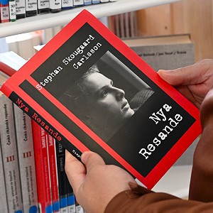 Händer håller boken 'Nya Resande' av Stephan Skougaard Carlsson med rött omslag och svartvitt porträtt, framför en bokhylla.