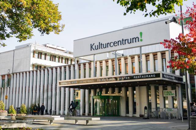Vit byggnad med stor text på fasaden där det står Kulturcentrum! Ovanför entrén står det texten: Folkbibliotek, konsthall, folkets hus, teater, restaurang. Ett träd med röda löv syns till höger i bild.