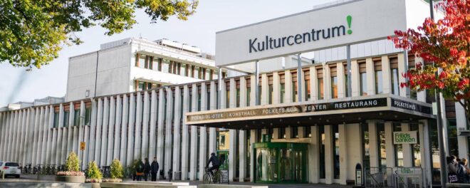 Vit byggnad med stor text på fasaden där det står Kulturcentrum! Ovanför entrén står det texten: Folkbibliotek, konsthall, folkets hus, teater, restaurang. Ett träd med röda löv syns till höger i bild.