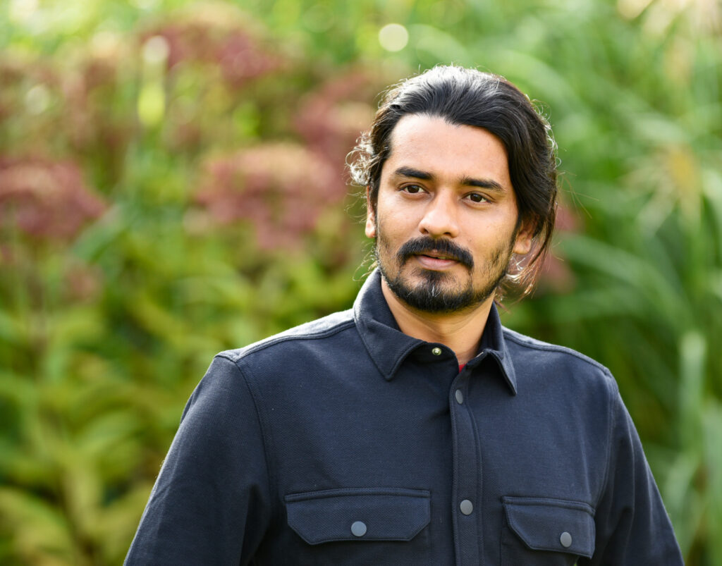 Porträtt på en man i svart skjorta som står framför en grönskande buske.