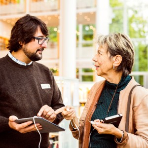 En äldre kvinna och en yngre manlig bibliotekarie i ett modernt bibliotek. De står tillsammans och tittar på en surfplatta som bibliotekarien håller. Kvinnan är klädd i en petrolblå stickad tröja och ljusrosa kofta, medan bibliotekarien bär en mörkgrå stickad tröja med namnbricka. I bakgrunden syns ljusa bokhyllor och stora fönster med utsikt mot grönska. Bilden förmedlar en hjälpsam och välkomnande stämning där bibliotekarien guidar besökaren i användning av digital teknik