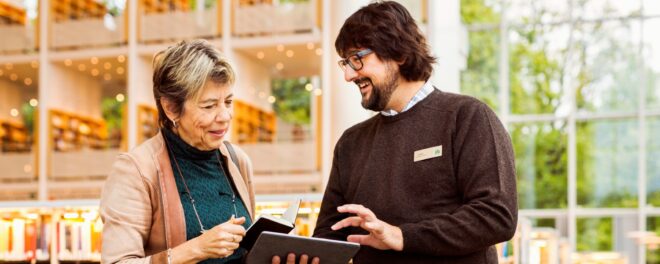En äldre kvinna och en yngre manlig bibliotekarie i ett modernt bibliotek. De står tillsammans och tittar på en surfplatta som bibliotekarien håller. Kvinnan är klädd i en petrolblå stickad tröja och ljusrosa kofta, medan bibliotekarien bär en mörkgrå stickad tröja med namnbricka. I bakgrunden syns ljusa bokhyllor och stora fönster med utsikt mot grönska. Bilden förmedlar en hjälpsam och välkomnande stämning där bibliotekarien guidar besökaren i användning av digital teknik.
