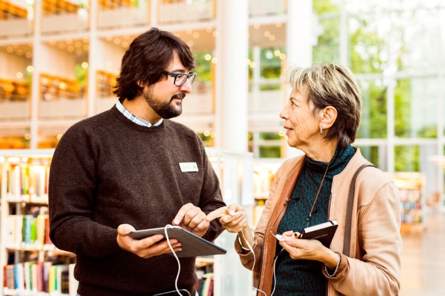 En äldre kvinna och en yngre manlig bibliotekarie i ett modernt bibliotek. De står tillsammans och tittar på en surfplatta som bibliotekarien håller. Kvinnan är klädd i en petrolblå stickad tröja och ljusrosa kofta, medan bibliotekarien bär en mörkgrå stickad tröja med namnbricka. I bakgrunden syns ljusa bokhyllor och stora fönster med utsikt mot grönska. Bilden förmedlar en hjälpsam och välkomnande stämning där bibliotekarien guidar besökaren i användning av digital teknik