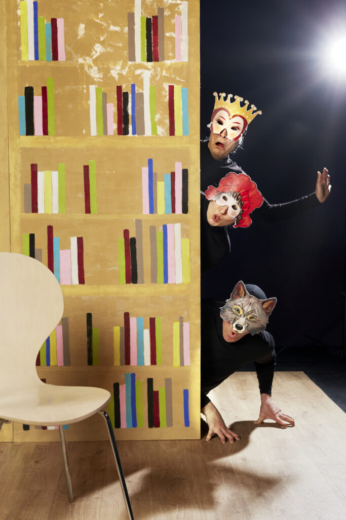 Tre människor i masker tittar fram med huvudena och halva kroppen, ifrån en vägg som är en målad bokhylla. Bakgrunden är svart och alla tre är klädda i svart. Personen som ses överst har en krona, och personen i mitten har en mask som är en röd mössa på huvudet, och den nedre en vargmask. Alla har förvånade ansiktsuttryck. 