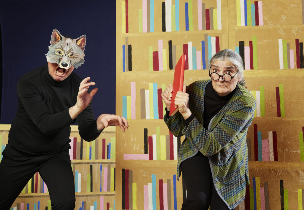 Två personer syns framför en vägg som är en målad bokhylla. Personen till vänster har en vargmask och personen till höger försöker skydda sig mot den andre genom att hålla upp en röd bok framför kroppen.