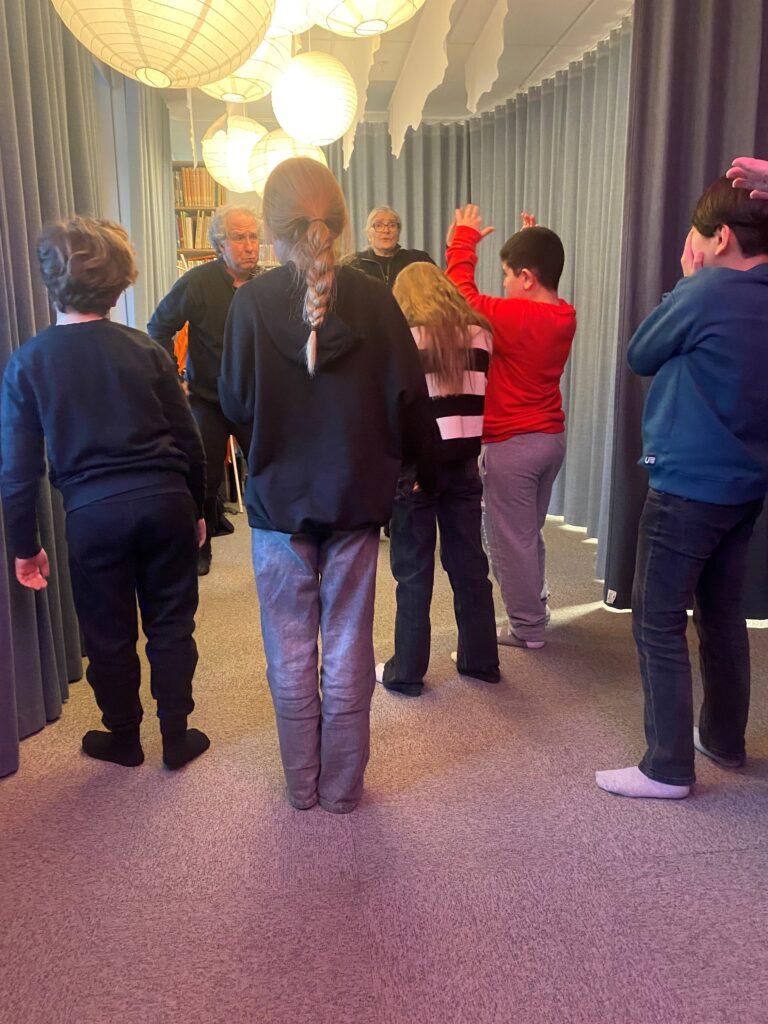 Två vuxna personer står längst fram och vända mot dem är fem barn, där vissa gör gester med händerna. 