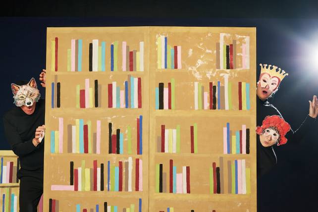 Tre figurer med masker på huvudena tittar fram ifrån en vägg som är målad som en bokhylla. Bakgrunden är svart och figurerna är klädda i svart. Den vänstra figuren har en vargmask, den översta högra har en mask med ögon och krona på huvudet och den nedersta till höger en mask med ögon och röd hatt.