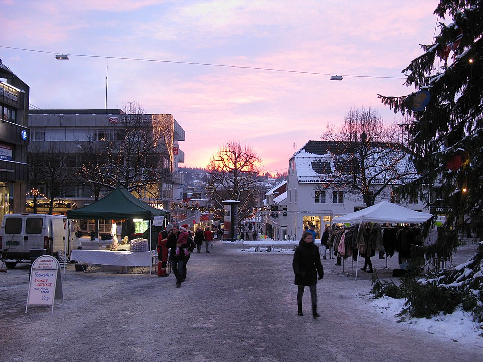 En stadsmiljö med snö på marken och en solnedgång i bakgrunden. En gran syns till höger i bild, några stånd som säljer kläder och annat, samt några människor som promenerar. Ljusslingor är upphängda i träd och över gatan.