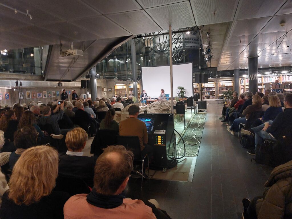 Publik med ryggen mot kameran, syns fylla ett rum. Längst fram syns två personer samtala på en scen med en vit filmduk bakom sig. Det är högt i tak.