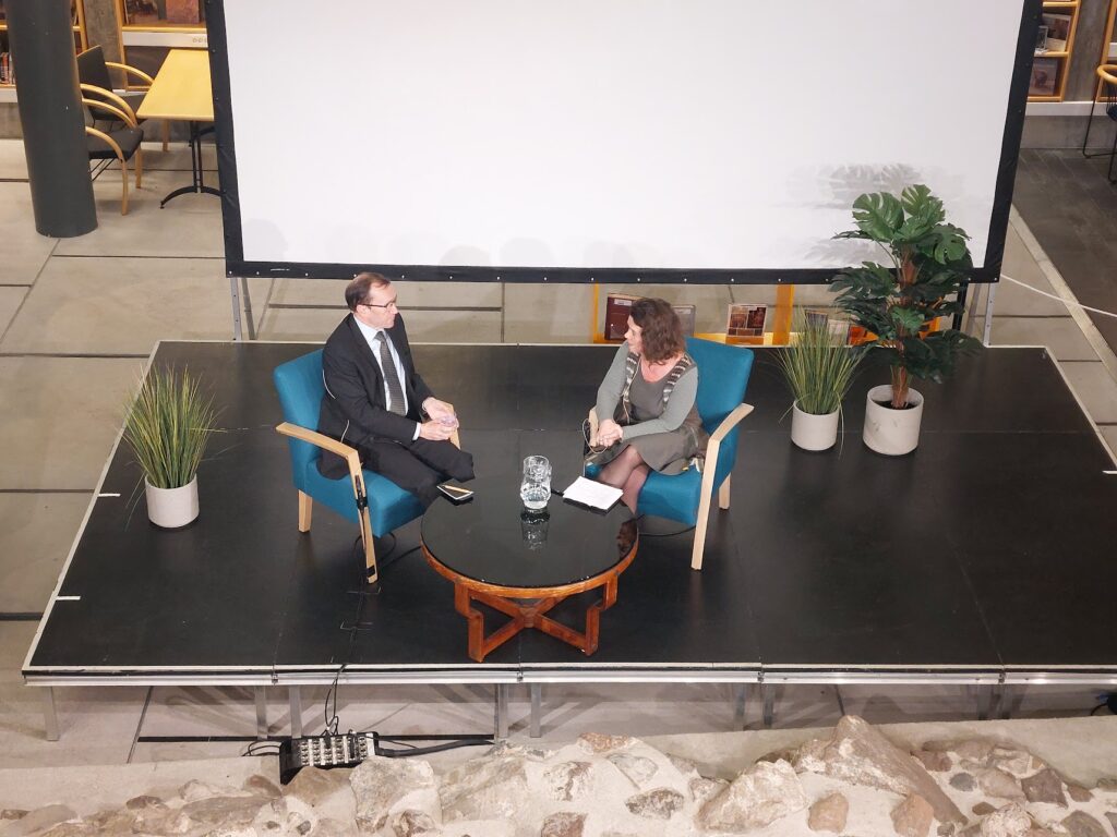 En man i kostym och slips sitter i en blå fåtölj och en kvinna i grön och grå kläder sitter i en fåtölj. De samtalar på en scen med några växte vid sidan om dem, och ett bord framför dem. Bakom dem syns en filmduk.