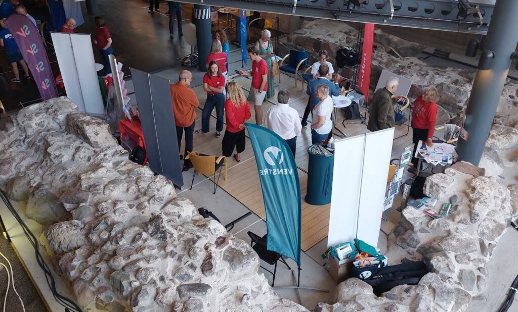 Ett tiotal människor står i små grupper eller två och två och samtalar, i ett rum med högt i tak. Runtom dem finns partiflaggor och rollups, det finns några stolar och bord med broschyrer mellan människorna. Som en inramning finns en stenmur.