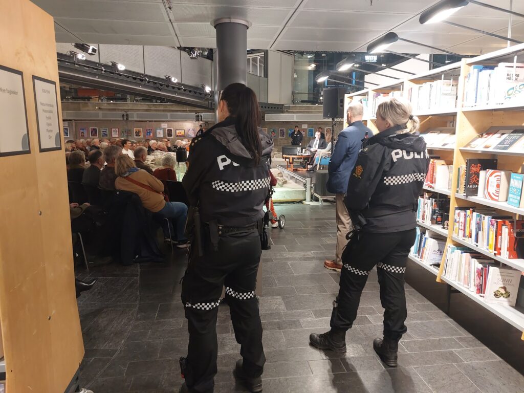 Två poliser står med ryggen emot kameran, intill en bokhylla och blickar ut över rummet som är fullt med människor som sitter och lyssnar. Längre fram skymtar en scen där två personer samtalar.