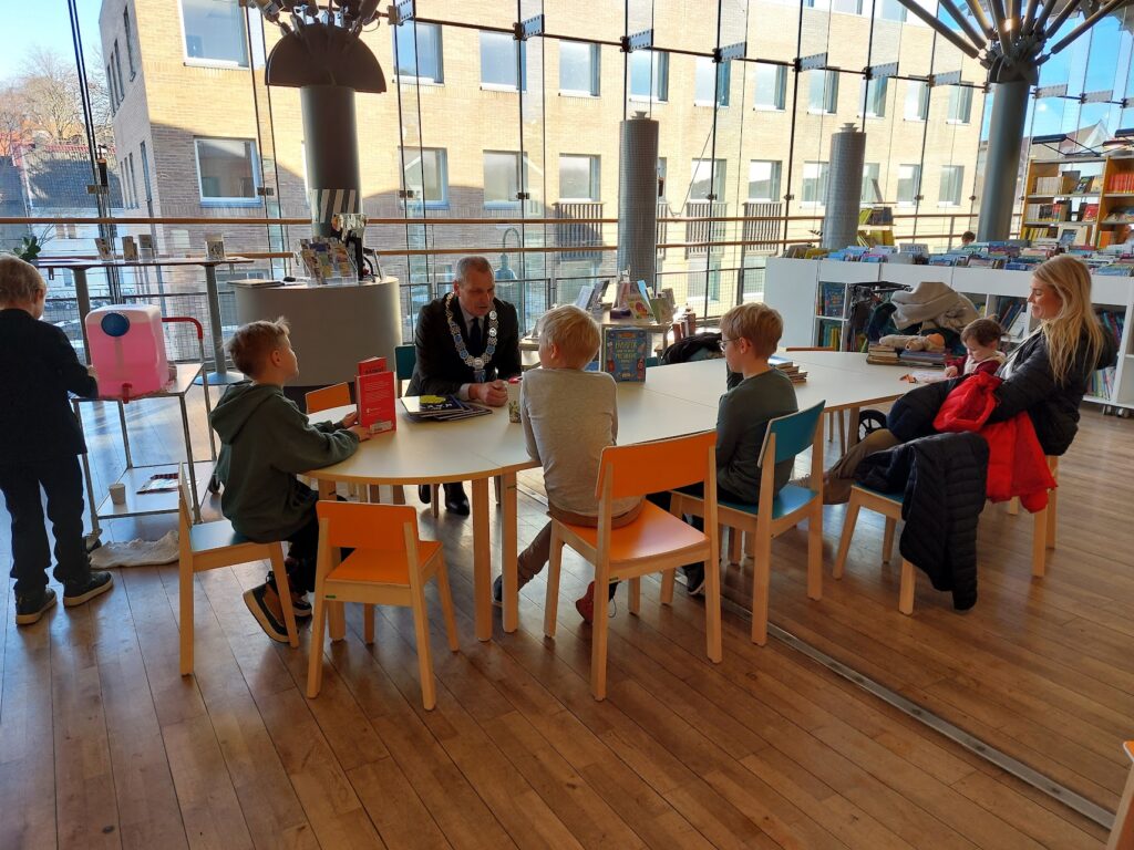 Runt ett bord sitter en man i kostym och med ett medaljerat halsband, och tre barn i tioårsåldern, samt en kvinna med en bebis famnen. Interiören är ett biblioteksrum med glasvägg ut mot ett annat hus. Ett barn syns ta dricka till vänster i bild.