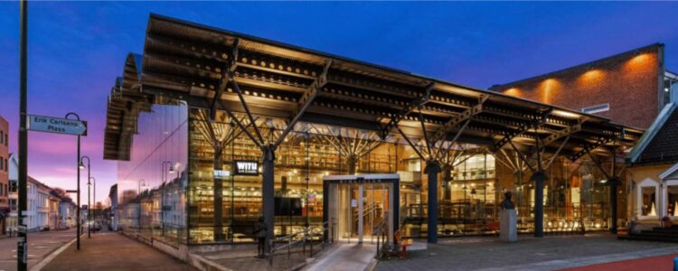 Byggnad med glasväggar i skymningsljus, med texten BIBLIOTEK ovanför ingången. Byggnaden är upplyst på insidan. På en skylt till vänster står det Erik Carlsens Plass och det sinns en väg till vänster i bild kantad av hus.