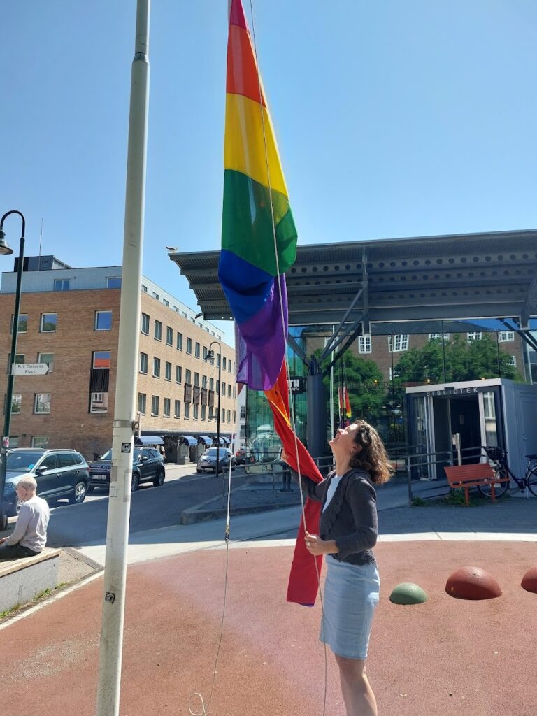 Ute på en öppen plats med blå himmer och framför en dörr där det står BIBLIOTEK, hissar en kvinna en flagga i gult, grönt, blått, lila och rött.