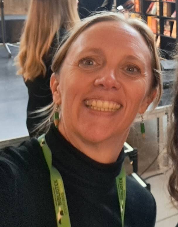 En leende kvinna med blont hår, svart tröja och grönt nyckelband.