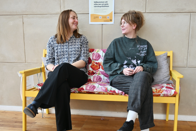 Två personer sitter på en gul bänk och tittar leende på varandra. Bakom dem syns en affisch där det står "Välkommen att sitta ner på gula vänskapsbänken".