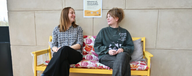 Två personer sitter på en gul bänk och tittar leende på varandra. Bakom dem syns en affisch där det står "Välkommen att sitta ner på gula vänskapsbänken".