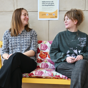 Två personer sitter på en gul bänk och tittar leende på varandra. Bakom dem syns en affisch där det står "Välkommen att sitta ner på gula vänskapsbänken".