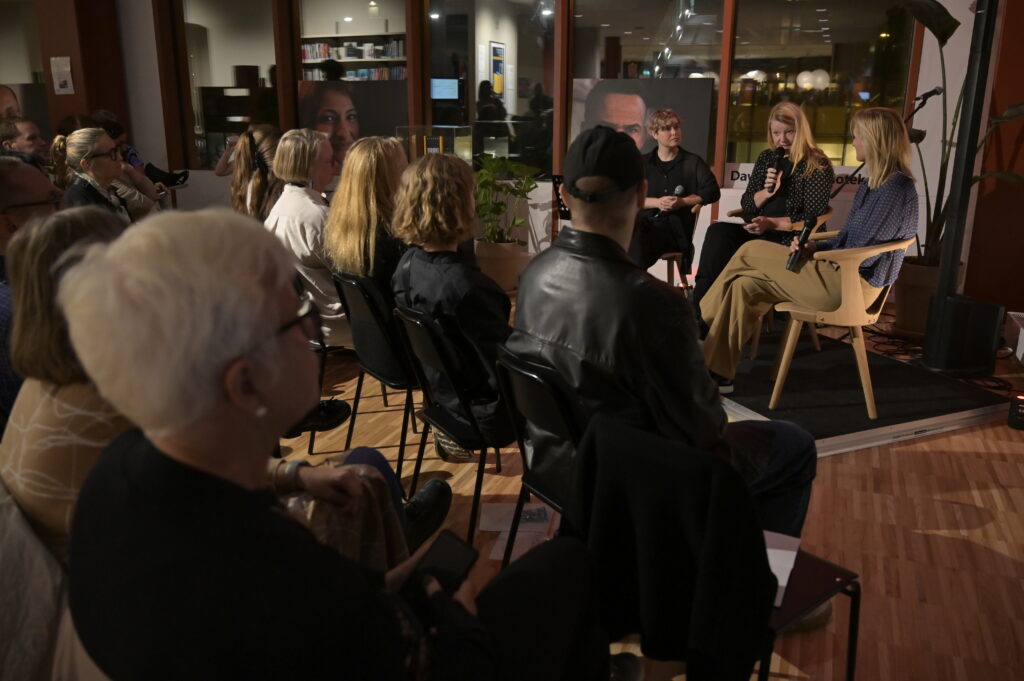 En sal med människor placerade som i en publik. De är vända mot en panel av tre personer, som alla sitter med en mikrofon i handen. I bakgrunden syns fönstren på biblioteken, det är ganska mörkt i lokalen.