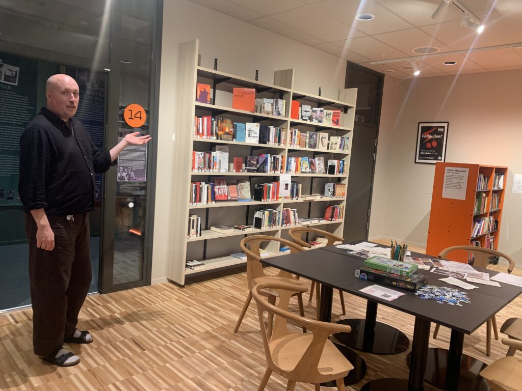 BIbliotekarie i svarta kläder visar med handen en lokal med bord, spel, kritor, pussel och bokhyllor runtomkring.