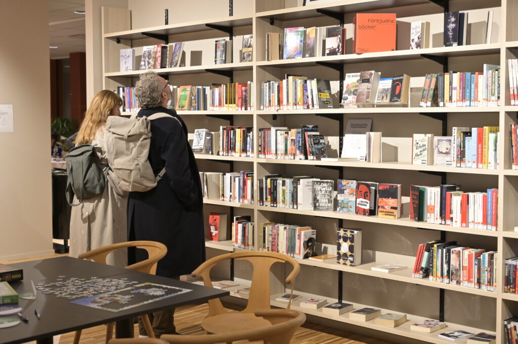 Ett biblioteksrum med en stor bokhylla. Framför den står två personer med ryggsäckar och läser på bokryggarna. Överst på hyllan är en orange skylt som säger "förbjudna böcker".