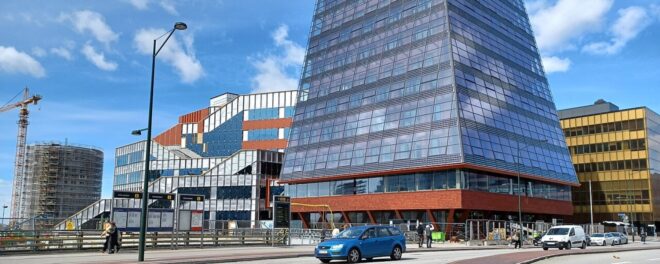 Den nedre delen av en tornliknande byggnad med glasfönster, syns till höger i bild. Den ligger i ett stadslandskap med en väg och en bil framför och andra byggnader runtomkring. Det finns en blå himmel bakom.