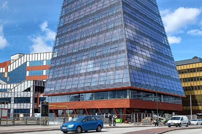 Den nedre delen av en tornliknande byggnad med glasfönster, syns i bild. Den ligger i ett stadslandskap med en väg och en bil framför och andra byggnader runtomkring. Det finns en blå himmel bakom.