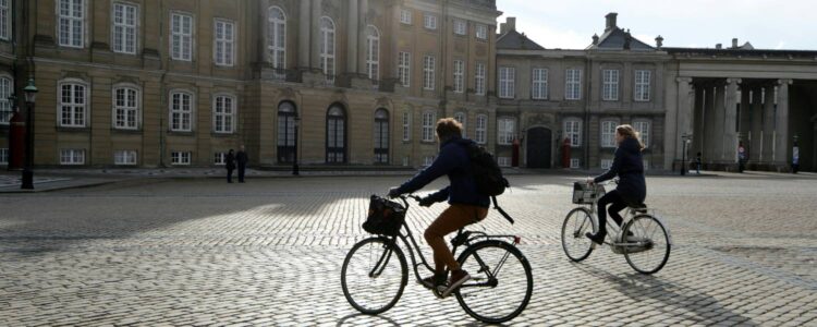 Två personer cyklar över ett torg med kullersten. Byggnader syns i bakgrunden och över dessa en lätt disig himmel.