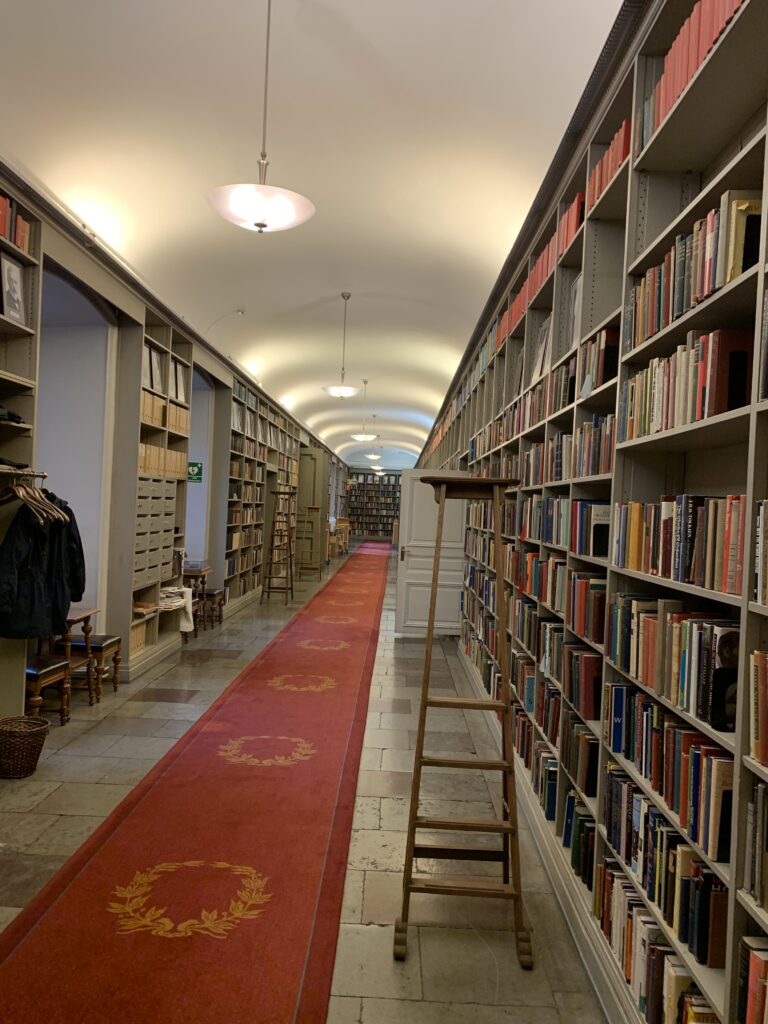 En korridor kantad av grå bokhyllor, fulla med böcker. En stege står till höger i bild vid en hylla. På golvet ligger en röd matta med Svenska Akademiens sigill i gult.