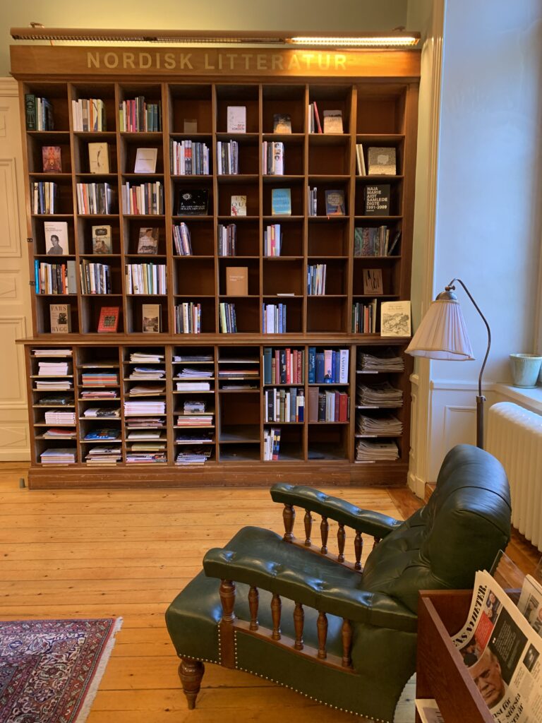 I bild syns en grön skinnfåtölj med stomme i trä, en golvlampa med skärm av tyg, och en bokhylla med olika fack. Främst i bild är ett tidningsställ med Dagens Nyheter. 