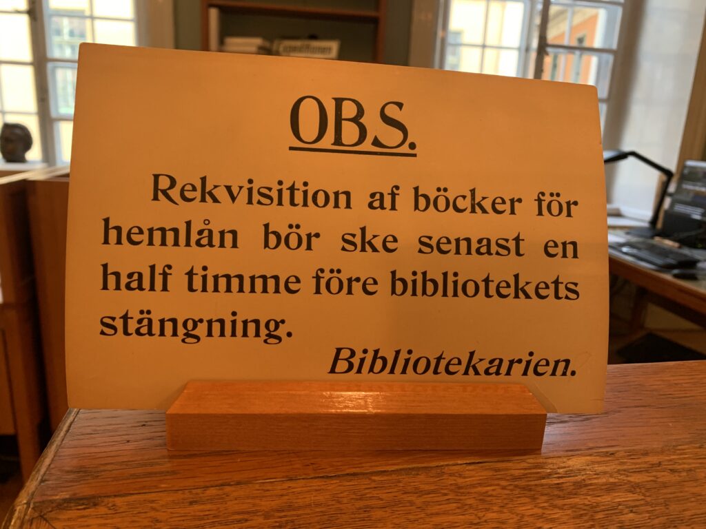 En gammeldags skylt där det står: OBS. Rekvisition af böcker för hemlån bör ske senast en half timme före bibliotekets stängning. / Bibliotekarien.