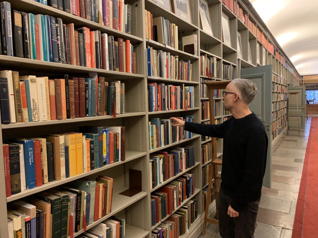 En bokhylla kantar en lång korridor, en man i svarta kläder och grått hår står bredvid och håller på att ta ut en bok. 