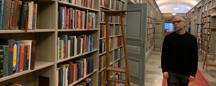 Ett rum kantat av bokhyllor. Till höger en man i svart tröja och glasögon som ser drömmande mot hyllorna.
